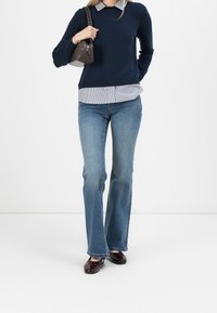 Pull marine porté sur une chemise à col rayée bleu et blanc, associé à un jean évasé et des chaussures bordeaux brillantes. Un petit sac à main sombre complète le look.