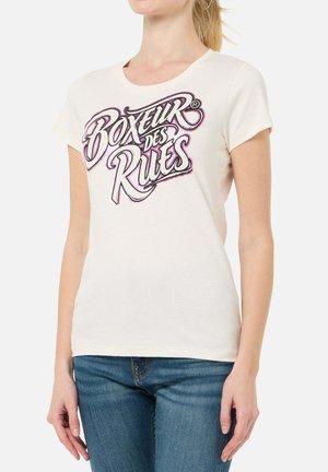 Frau trägt ein weißes Kurzarm-T-Shirt mit dem Text „Boxeur des Rues“ in fetter, stilisierter Schrift, kombiniert mit blauen Jeans.
