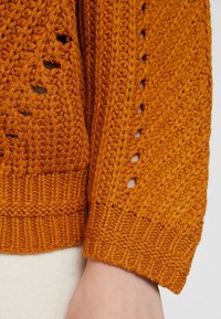 Strickpullover in einem satten Orangeton, mit Lochmuster-Designs und gerippten Bündchen. Die Textur ist dick und kuschelig mit einem ausgeprägten Muster.