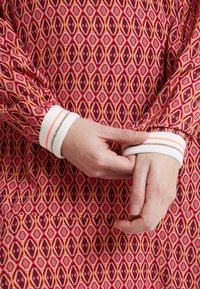Robe rouge à motifs avec des formes en losange en noir, pêche et jaune. Les poignets blancs présentent des rayures alternées de couleur. Des mains ajustent la manche.