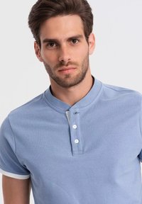 Světle modré polo tričko s límečkem na knoflíky, krátkými rukávy s bílými akcenty a strukturovanou látkou. Detailní pohled na tričko.