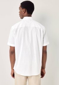 Uomo visto da dietro che indossa una camicia bianca a maniche corte con bottoni e pantaloni beige su uno sfondo chiaro e uniforme.