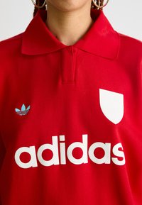 Maglietta polo rossa in cotone, con logo bianco "adidas" e logo a tre strisce blu sul petto, dotata di un design classico del colletto.
