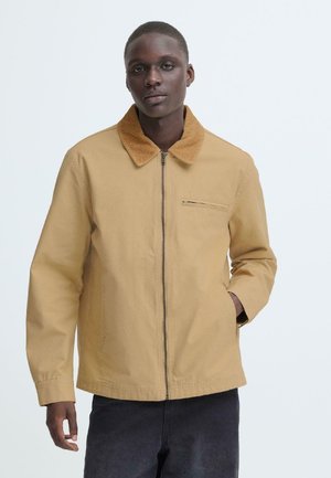 Man draagt een beige jas met rits en een corduroy kraag en donkere broek, staat tegen een effen lichte achtergrond.