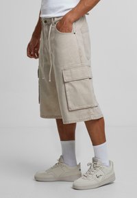 Beige cargo šortky s dvěma velkými bočními kapsami, stahovací pas a volný střih. Pár s bílými teniskami a ponožkami po kotníky.