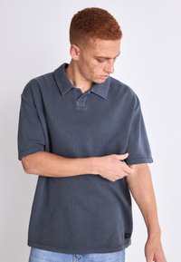 Tmavošedé polo tričko s textúrovanou tkaninou, krátkymi rukávmi a gombíkovým golierom. Obsahuje malé logo na dolnom ľavom rohu.