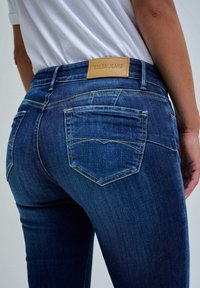 Persona que lleva vaqueros de mezclilla azul oscuro con bolsillos traseros y una etiqueta de cuero con la marca "Salsa Jeans."