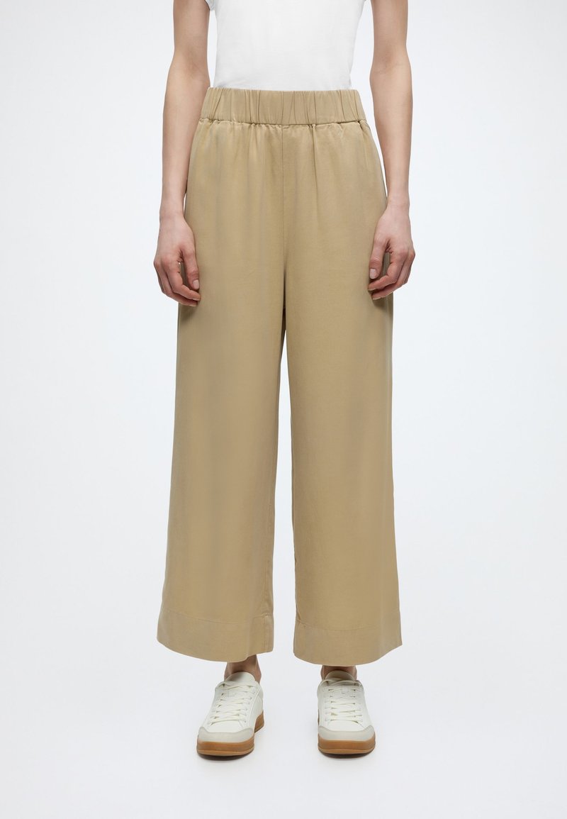 Άτομο που φοράει μπεζ παντελόνι με φαρδιά πόδια και ελαστική μέση, λευκά αθλητικά παπούτσια και λευκή μπλούζα σε απλό φόντο.