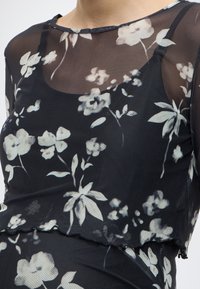 Μαύρο διαφανές μπλουζάκι με λευκό λουλουδάτο σχέδιο, με στρογγυλή λαιμόκοψη, μακριά μανίκια και κυματιστό τελείωμα. Ελαφρύ, υφασμάτινο ύφασμα με υφή.