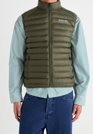 Person iført en grøn vatteret Red Bull Racing vest over en grøn og hvidstribet skjorte og blå jeans, stående med afslappede hænder.