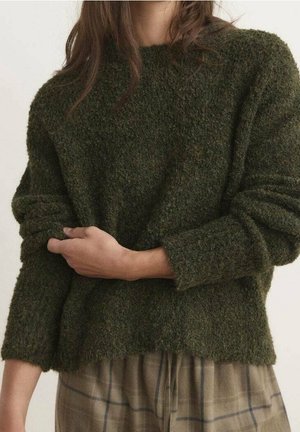 Kobieta w ciemnozielonym swetrze o teksturowanej powierzchni z podwiniętymi rękawami i w kratę spodniach, trzymająca jedną rękę drugą.