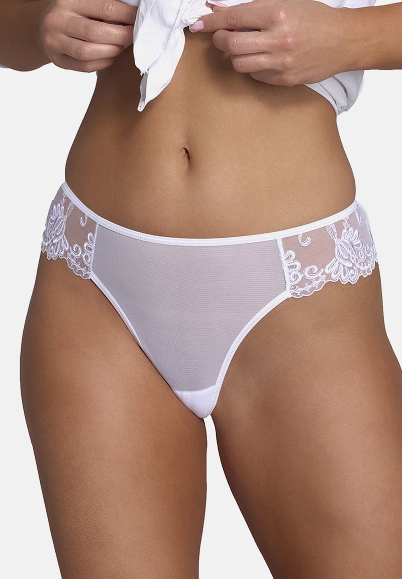 Vrouw die witte kanten en gaas bikini-achtige onderbroek aanpast, zichtbaar middenrif en bovenbenen, tegen een effen lichte achtergrond.
