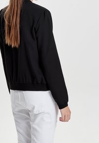 Giacca bomber nera con zip, maniche lunghe, polsini arricciati e vita elastica. Indossata con jeans bianchi, mette in mostra un design elegante e minimalista.