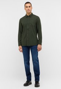 Donkergroene langemouwen shirt met een knoopsluiting aan de voorkant en een enkele borstzak, gecombineerd met blauwe spijkerbroeken en zwarte schoenen.