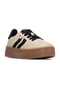 Zapatillas sintéticas beige con acentos de rayas negras, puntera redonda, suela de goma gruesa color tostado y cordones negros. Textura suave con detalle del logo.