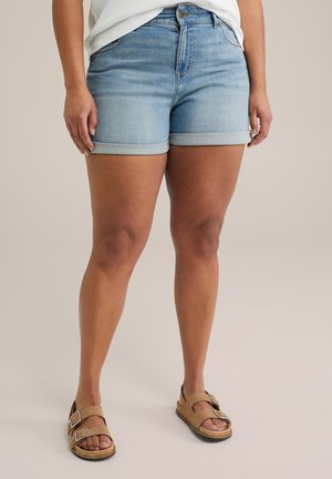 Person iført lyseblå opkørte denimbukser, hvid top og tan dobbelt-spænde sandaler stående på neutral baggrund.