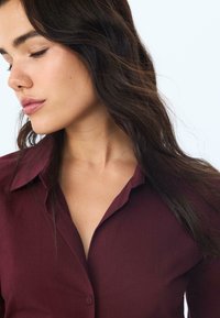 Burgunderfarvet skjorte med krave, der har en glat tekstur og en let glans. Skjorten vises mod en lys baggrund.