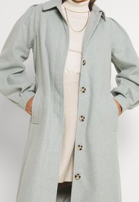 Manteau en laine vert clair avec un col pointu, des manches longues, deux poches latérales et de grands boutons bruns sur le devant. Texture lisse et coupe structurée.