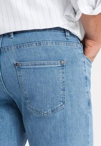 Osoba v světle modrých džínech a bílé pruhované košili, s jednou rukou opřenou o přední kapsu džínů.