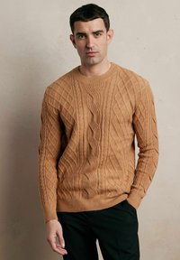 Brązowy sweter w splot brązowy z teksturowanymi wzorami, okrągłym dekoltem i ściągaczami, stylizowany z czarnymi spodniami.