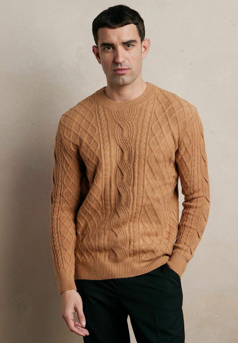 Brązowy sweter w splot brązowy z teksturowanymi wzorami, okrągłym dekoltem i ściągaczami, stylizowany z czarnymi spodniami.