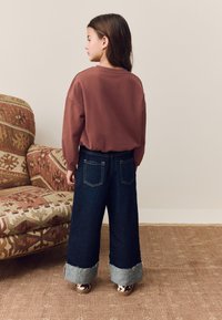 Viininpunainen ylisuurikokoinen collegepaita, tumman siniset leveälahkeiset farkut, joiden hihansuut ovat rispaantuneet, ja kuvioidut kengät. Taustalla ruskea kuvioitu sohva.