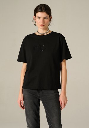T-shirt in cotone nero con vestibilità ampia, maniche corte e logo "MC" in paillettes sulla parte anteriore. Abbinato a jeans scuri.