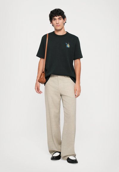 Tânăr �îmbrăcat cu un tricou negru cu grafică, pantaloni largi bej, loafers alb-negru și o geantă de umăr maro, stând în fața unui fundal alb.