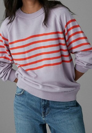 Persona con suéter lavanda con cinco rayas horizontales rojas en el pecho y jeans azules, con las manos detrás de la espalda.