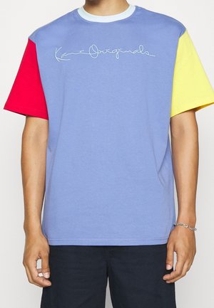 Man draagt een T-shirt met kleurvlakken: een rode linkermouw, gele rechtermouw, blauwe romp en een wit logo in sierletters op de borst.