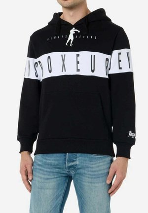 Mann trägt schwarzen Hoodie mit weißem horizontalem Streifen mit schwarzen Buchstaben und Box-Silhouette, kombiniert mit blauen Jeans.
