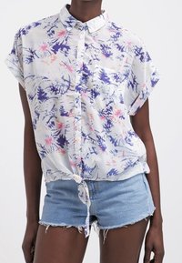 Biała koszula z kwiatowym wzorem w fioletowe i różowe motywy. Krótkie rękawy i przewiązana w talii. W połączeniu z postrzępionymi szortami dżinsowymi.