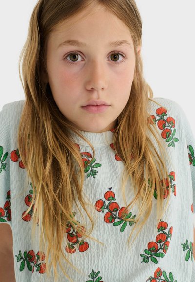 Jeune fille aux longs cheveux blonds portant une chemise blanche avec des motifs de tomates rouges, regardant droit devant elle avec une expression neutre.