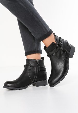 Zwarte enkellaarzen met een zijrits en decoratieve band, gedragen met opgerolde donkere jeans over elkaar geslagen tegen een witte achtergrond.