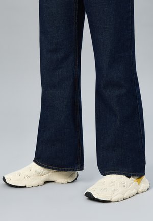 Jeans a zampa di colore blu scuro indossati con sneakers slip-on bianche con texture, caratterizzate da dettagli gialli e suola nera.