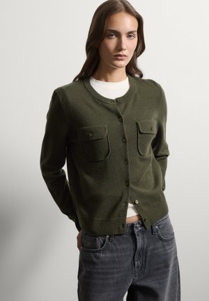 Giovane donna che indossa un cardigan verde scuro abbottonato con tasche frontali sopra una maglia bianca e jeans grigi slavati su uno sfondo semplice.