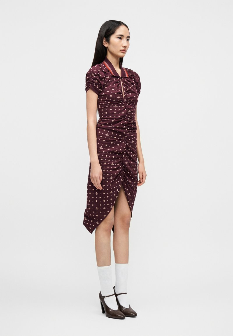 Frau in einem burgunderroten, gerafften Kleid mit floralem Muster, kurzen Puffärmeln, weißen Socken und braunen Mary-Jane-Pumps, die vor weißem Hintergrund steht.