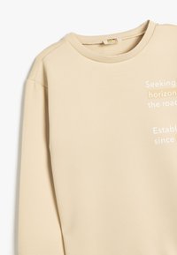 Beige sweatshirt med rund halsudskæring, prydet med hvid tekst på forsiden. Glat tekstur og afslappet pasform, med raglanærmer for komfort.
