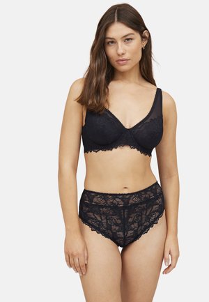 Kvinde med langt brunt hår iført en sort blonde-bh og matchende højtaljede blonde-trusser, stående mod en hvid baggrund.