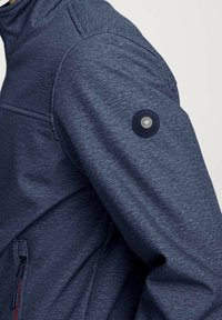 Marineblauwe softshell jas met een gestructureerd oppervlak, getailleerde pasvorm en een knoopdetail op de mouw. Voorzien van een ritszak aan de zijkant.