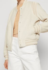 Veste bomber couleur crème en tissu texturé, dotée de détails côtelés et de boutons-pression, associée à un haut beige et un pantalon blanc.