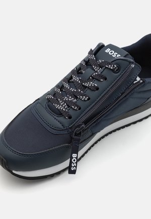 Tmavě modrá teniska se směsí texturovaného nylonu a hladké kůže, vybavená šedými tkaničkami, bočním zipem a bílou podrážkou.