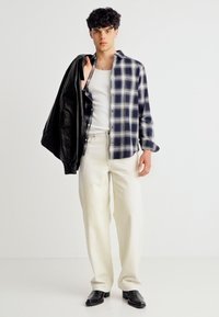 Νέος άνδρας με λευκό αμάνικο μπλουζάκι, μπλε και λευκό καρό πουκάμισο, εκρού παντελόνι, μαύρα μποτάκια, κρατώντας μαύρο δερμάτινο μπουφάν στον ώμο.