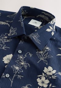 Kurzärmeliges Hemd in Marineblau mit strukturierter Stoffmusterung und weißem Blumenmuster. Verfügt über einen strukturierten Kragen und zwei Knöpfe vorne.