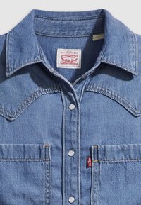 Jasnoniebieska koszula jeansowa z ostrym kołnierzem, zapinana z przodu na zatrzaski, z kieszeniami na piersi i czerwoną metką Levi's po boku, ozdobiona kontrastującymi żółtymi szwami.