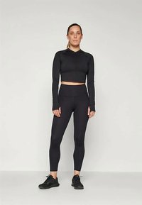 Svart treningssett bestående av en cropped langermet topp og høy midje tights, laget av et glatt og elastisk stoff, sammen med svarte joggesko.