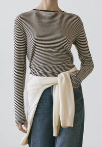 Gestreiftes Langarmshirt in Beige und Schwarz mit figurbetontem Schnitt. Kombiniert mit einem hellen Pullover, der um die Taille gebunden ist, und blauen Jeans aus Denim.