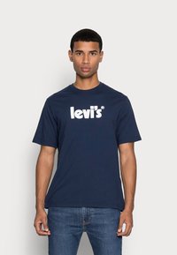 Navy blue puuvillane T-särk, millel on valge "Levi's" logo, ümar kaelus ja lühikesed varrukad, koos teksapükstega. Sile tekstuur.