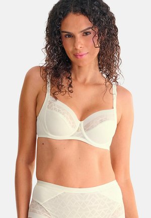 Femme aux cheveux bouclés portant un soutien-gorge blanc à armatures et une culotte transparente à taille haute assortie, face à l'avant.