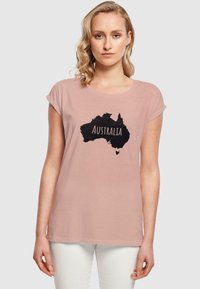 Rosa t-skjorte med korte ermer som har en svart kontur av Australia med ordet "AUSTRALIA" i fet skrift. Myk tekstur av stoffet.